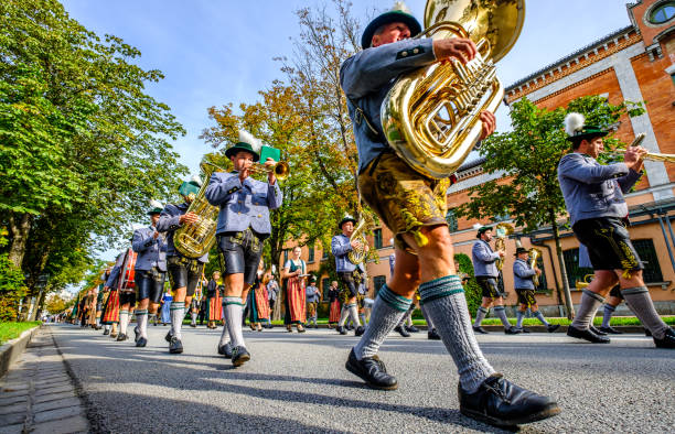 Munich, Germany - September 21, 2025:   Vibrant street parade featuring musicians in red jackets, lederhosen and dirndls performing with saxophones and clarinets under bright daylight. A festive, cultural moment with crowds and urban backdrop.
