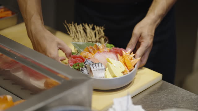 Sashimi chef arranging sweet egg roll on a sashimi plate full of colorful sliced raw fish and seafood on kitchen counter and placing it on serving counter with beautiful presentation at a sushi restaurant.