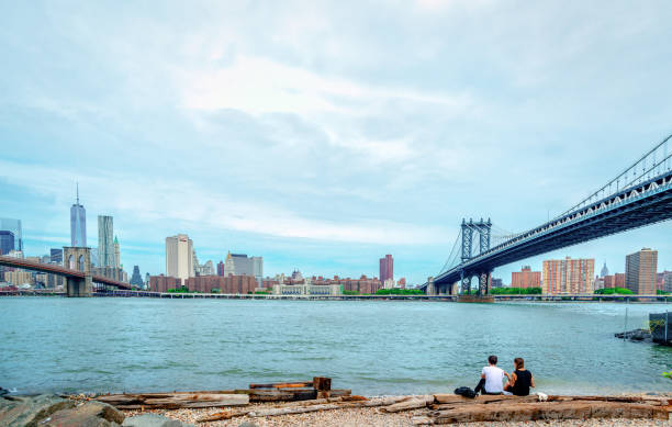 jeune couple au bord de l’east river avec la ligne d’horizon de manhattan en arrière-plan. - brooklyn bridge national landmark brooklyn bridge park dumbo photos et images de collection