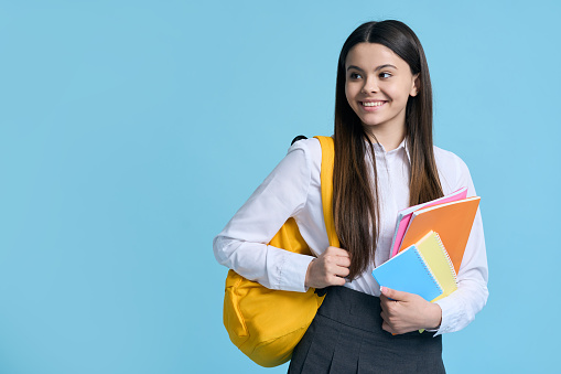 Happy Caucasian teenager, schoolgirl with yellow backpack and textbooks, smiles looking aside, isolated on blue color background. People. Education. Back to school on new semester of academic year