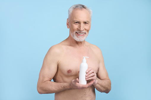 Waist-up portrait Caucasian senior man holding white bottle with moisturizing nourishing body lotion, smiling looking at camera, posing with torso naked, isolated on blue background. Skin care concept