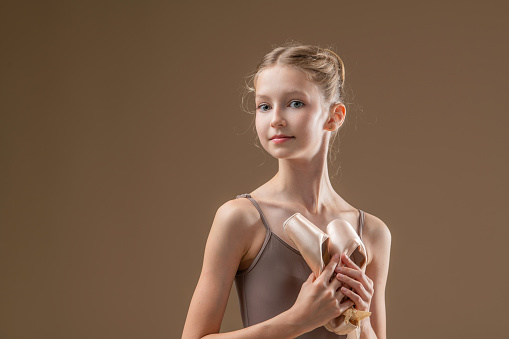 Portrait of beautiful young girl professional ballerina student with pointe shoes in a leotard on a light beige background.