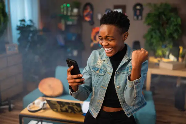 Photo of Happy african american teen girl lady woman with short hair using cell phone holding in hands smiling celebrating raising fists