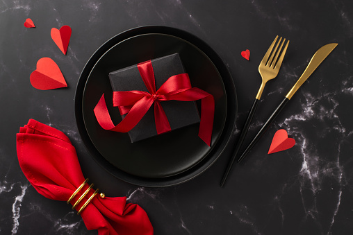 Dine-in restaurant on Valentine's Day theme. Top-view shot of beautiful giftbox in plate, hearts, elegant tableware, red napkin with ring on romantic black marble table setting, perfect for ad