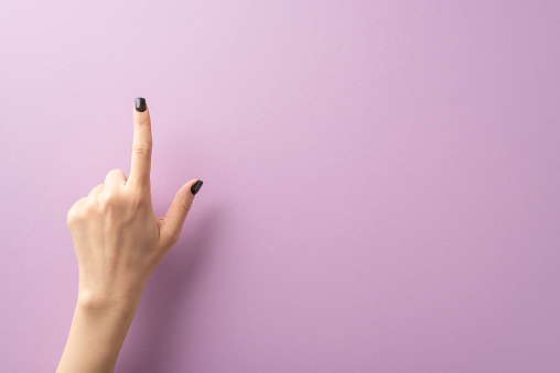First person top view showcases fashionable young lady's hand, donning chic black nail polish, shaping choosing sign with her index finger. Purple background offers space for text or advertisements
