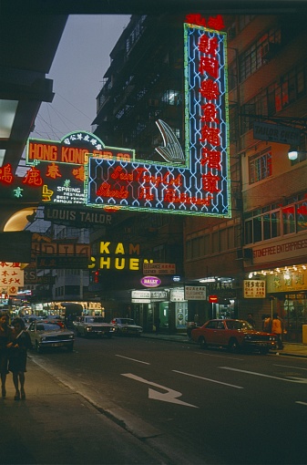 British Hong Kong, China, 1979. Commercial street with neon signs in British Hong Kong. Furthermore: pedestrians, vehicles, shops and buildings.