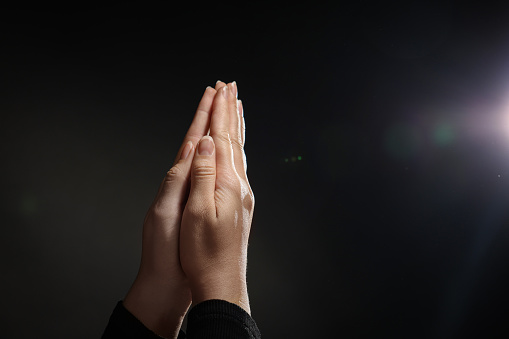 Woman holding hands clasped while praying against light in darkness, closeup. Space for text