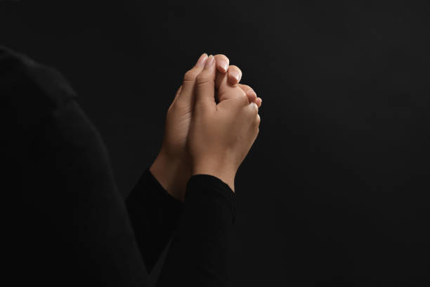 istock Woman holding hands clasped while praying against black background, closeup. Space for text 1728400984