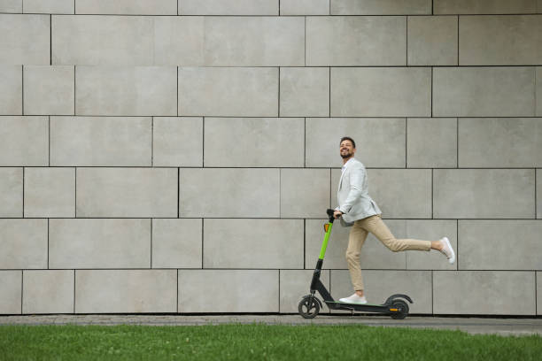 istock Businessman riding modern kick scooter near grey stone wall outdoors, space for text 1728473972