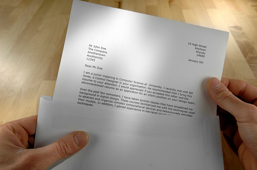 A point of view – POV – image of a caucasian man’s hands pulling a generic printed job application letter from a white envelope over a wooden desk. All information in the letter is fictional. 