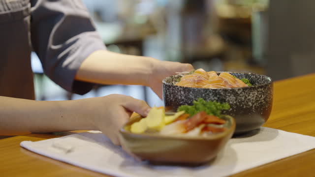 Close-up shot of Chirashi donburi and a plate of sliced Hokkigai or surf clam sashimi and tamagoyaki on kitchen counter while a waiter picking them up for serving.