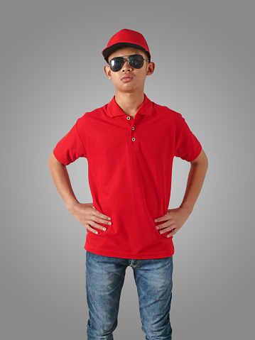 Young Asian teenage boy wearing red collared shirt, hat and sunglasses standing with hands on hip over grey background, t-shirt template mock up