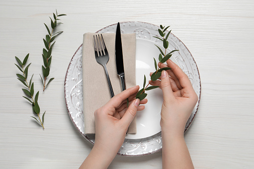 Woman adjusting stylish table setting with eucalyptus leaves on white wooden background, closeup