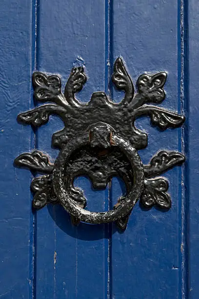 An old blue door and door knocker, painted black many times and rusty underneath. In strong sunlight.