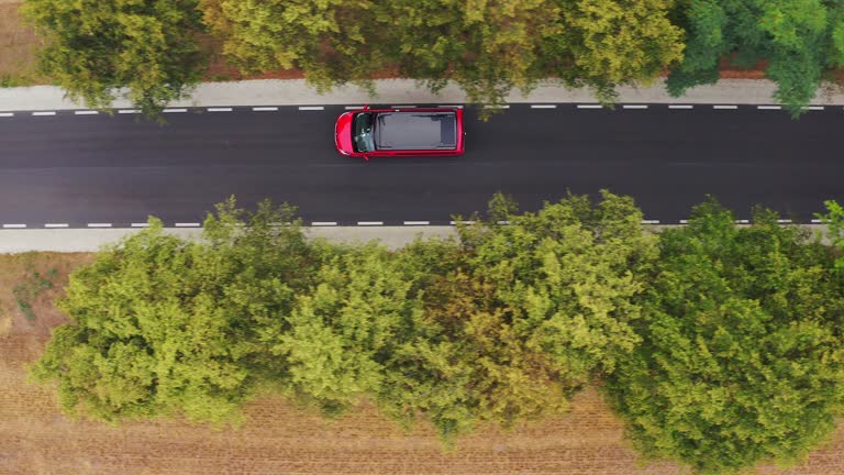 Drone top down asymmetrical follow shot of red minivan camper with black roof with solar panels driving along a narrow unmarked road in alley of trees.