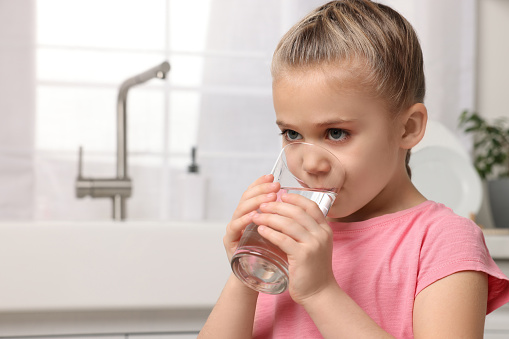 Cute little girl drinking fresh water from glass in kitchen