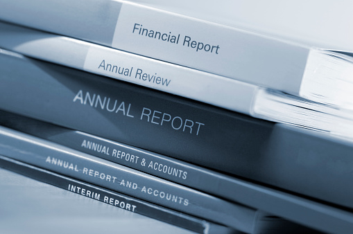 A blue toned close up image of a loosely arranged stack of thick generic business annual financial annual and accounts.  The reports are arranged on a wooden table and shot with a shallow depth of field. 
