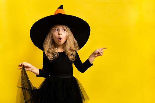 a little blonde girl in a black witch costume on a yellow background shows her hands in both directions to the place for the text, a happy and cheerful child.