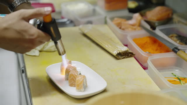 High angle view of chef's slicing pieces of salmon and wrapping it on a maki sushi and using bamboo mat to roll it tightly in order to make an inside out salmon sushi roll and using blow torch to grill the salmon at a sushi restaurant.