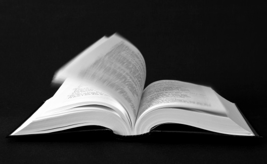 Black and white picture of an open book on black background whose pages are flipped by the wind