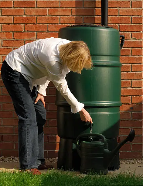Blonde woman filling a watering can from a rainwater butt/storage tank.