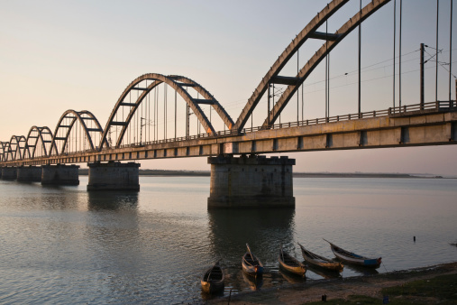 Fishing boats anchored on the shore of the Godavari River at sunset with railroad bridge in the background.