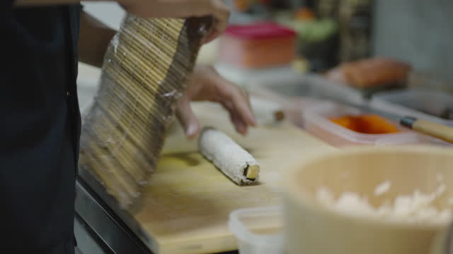 Side view of chef's hands putting pieces of sweet tamagoyaki, fresh carrot and cucumber on vinegared rice and seaweed on bamboo mat and rolling tightly to make a maki sushi on kitchen counter at a sushi restaurant.