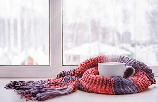 A cup of hot tea wrapped in a knitted terra cotta color scarf stands on the windowsill on a cold winter day
