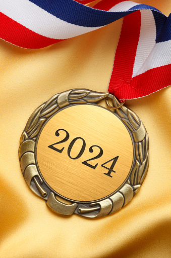 A gold medal engraved with 2024 on it.  A red, white, and blue ribbon is attached to the medal that rests on a piece of gold satin fabric.