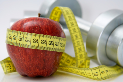 Close-up of an apple with a measuring tape around it and chrome dumbbells at the background. Shallow depth of field.