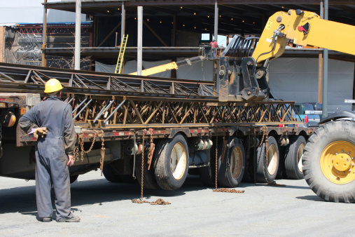 Steel girders are unloaded from a flatbed trailer truck on a construction site with a large fork lift.