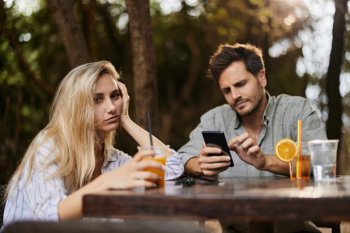 Young man using cell phone in an outdoor café while his girlfriend is feeling bored.
