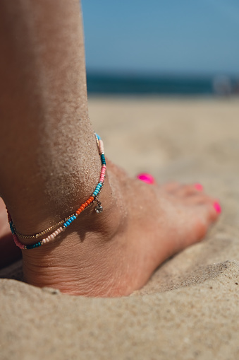 Girl wearing colorful ankle bracelet with pink pedicure. Sitting on sandy beach. Tropical Beach travel concept. Female summer jewelry. Holiday vacation