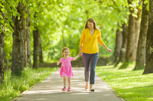 Happy smiling little daughter and young adult mother running on sidewalk at park of tree alley. Spending time together in beautiful warm sunny summer day. Front view.