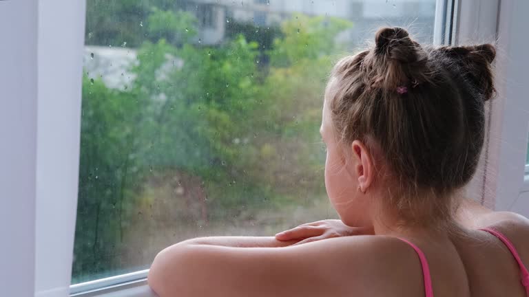 Sad little upset girl looks out the window. The child is bored near the window.