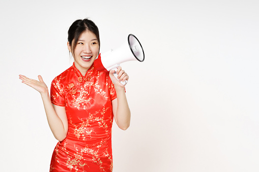 Asian woman wearing traditional cheongsam qipao dress with gesture holding megaphone isolated on white background. Happy Chinese new year.