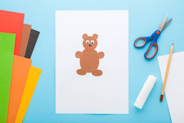 istock Brown teddy bear created from application paper on white sheet. Scissors, glue stick, pencil and colorful paper on light blue table background. Pastel color. Closeup. Top down view. 1729097366