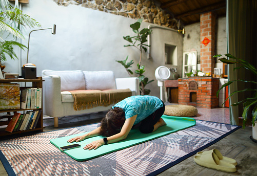 An elderly Asian woman is attending her morning yoga class while watching a video on her phone, lying face down on her yoga mat doing Balasana yoga pose