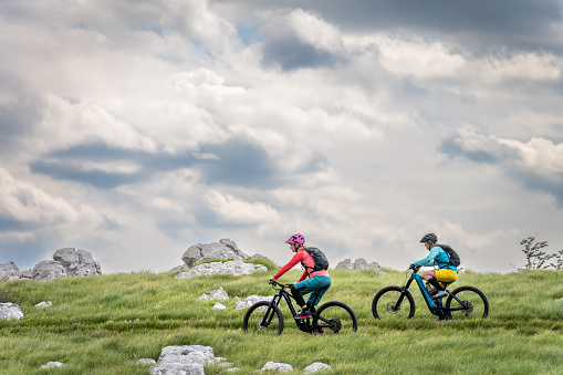 Two women riding electric mountain bikes together at green meadows and cloudscape scenery wide shot side view