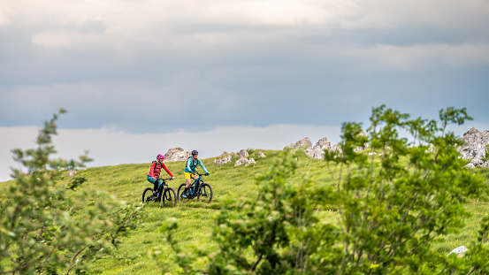 Two women riding electric mountain bikes together at green meadows and cloudscape scenery wide shot side view