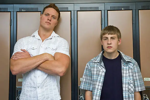 An intimidating guy stands next to a little guy.  Could be a bully or a bodyguard.  Standing in front of lockers.
