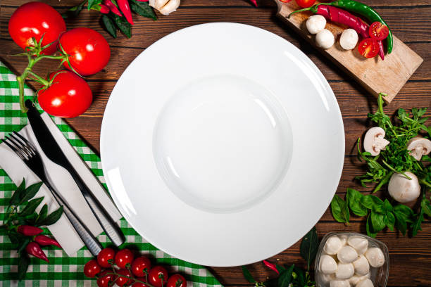 istock Round pasta plate decorated with tomatoes, mushrooms, spinach, basil and spices. Dish on a white square plate with cutlery on a wooden table, top view 1729152003