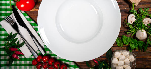 istock Round plate decorated with tomatoes, mushrooms, spinach, basil and spices. Dish on a white square plate with cutlery on a wooden table, top view 1729152038