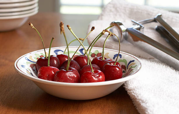 cherries in a bowl long stemmed red cherries in a bowl surrounded by a cherry pitter and some bowls set on a wooden surface. pitter stock pictures, royalty-free photos & images