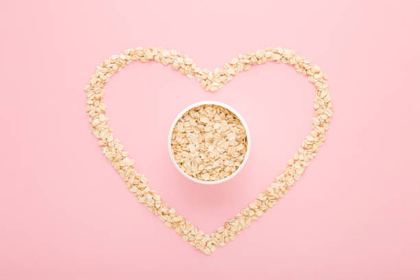 volle weiße schüssel und herzform aus trocken gerolltem hafer auf hellrosa tischhintergrund. pastellfarbe. ich liebe gesundes essen. closeup. draufsicht nach unten. - oatmeal oat heart shape rolled oats stock-fotos und bilder