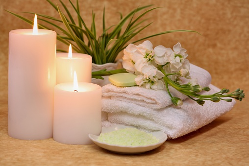 Tranquil spa still life scene with three burning white candles, folded towels, flowers, plant and light green bath salt in white dish. Horizontal image. For more spa images click below.