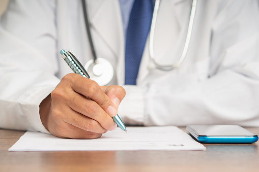 A doctor is holding a pen and writing on a medical prescription while sitting in the hospital. Medical personnel, medicine, and science concept. Quality medical services