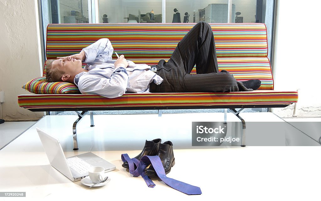 tired businessman lying on multi colored sofa tired businessman with hand on head lying down on multi colored sofa is looking at his mobile phone and took off his necktie and shoes Adult Stock Photo