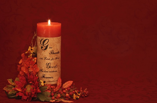 Burning autumn Candle with scripture from 1 Chronicles 16:34 on burgundy brocade and autumn colored flowers around candle. Horizontal Christian thanksgiving image with copy space.