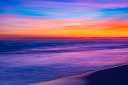 A stunning, long exposure sunset at the beach in Hikkaduwa. The sky is a gradient of pink, orange, and blue colors, creating a mesmerizing effect. The ocean is a deep blue color with white waves crashing onto the shore. The sand is a dark color and appears to be wet. The horizon line is visible in the distance.